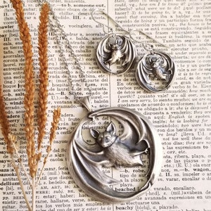 May include: A silver pendant featuring a bat with outstretched wings. The bat is detailed with a textured surface and has a small, round face. The pendant is hanging from a silver chain and is resting on a page of a book with text.