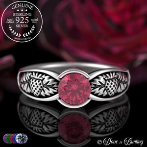 Puede incluir: Un anillo de plata de ley con una gema redonda de color rojo brillante. La banda presenta un diseño detallado de fresas y hojas. El anillo está marcado con "925 SILVER" y "MADE IN THE USA".