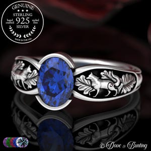 May include: A sterling silver ring with a large oval-cut blue gemstone. The band features a detailed design of foxes and foliage. The ring is marked "925 Silver" and "Made in the USA".