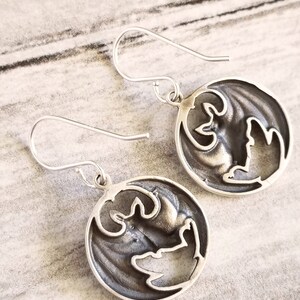 May include: A pair of silver earrings with a circular design featuring a cutout of a bat. The earrings have a simple hook closure.