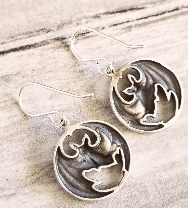 May include: A pair of silver earrings with a circular design featuring a cutout of a bat. The earrings have a simple hook closure.
