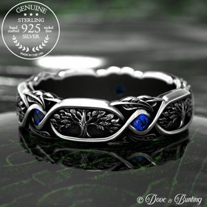 May include: A sterling silver ring with a detailed tree of life design and blue gemstone accents. The ring is marked "925 Silver" and "Made in the USA". The ring has a dark interior and a polished exterior.