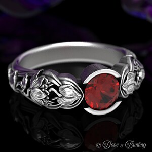 May include: A silver ring with a red gemstone set in a floral design. The ring is made of sterling silver and features a delicate floral pattern on the band.