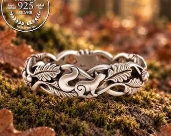 Puede incluir: Un anillo de plata de ley con un diseño de banda continua que presenta ardillas, hojas de roble y bellotas esculpidas. El metal tiene un acabado pulido con áreas empotradas oscurecidas para resaltar los detalles del motivo forestal.