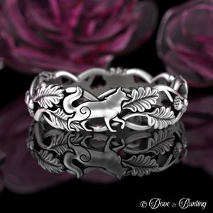 May include: A silver ring with a wolf design surrounded by leaves and acorns. The ring is on a black surface with a purple background.