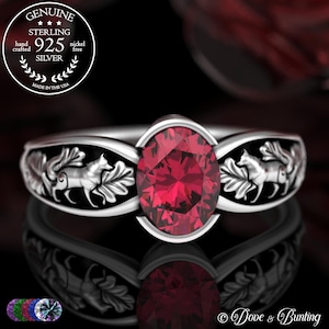 May include: A sterling silver ring with a large oval-cut red gemstone. The band features a detailed design of foxes and leaves. The ring is hand-crafted and nickel-free, made in the USA.