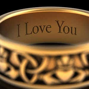 May include: Close-up of a gold ring with the inscription "I Love You" engraved on the inside. The ring has a Celtic knot design on the outer band, showcasing intricate details and a polished finish. The ring is set against a black background.
