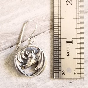 May include: Silver bat earrings with a circular design. The bat is in flight with its wings spread out. The earrings are about 1 inch in diameter.