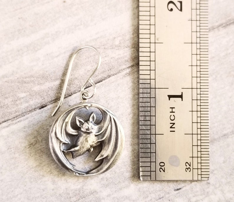 May include: Silver bat earrings with a circular design. The bat is in flight with its wings spread out. The earrings are about 1 inch in diameter.