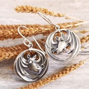 May include: Silver earrings with a bat design. The bats are in a circular shape with their wings spread out. The earrings are hanging from a silver chain.