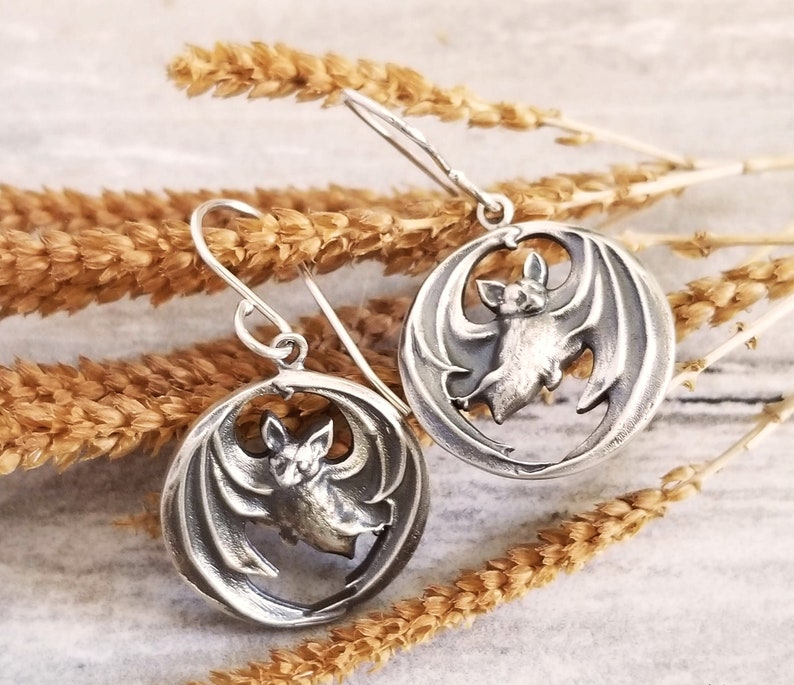 May include: Silver earrings with a bat design. The bats are in a circular shape with their wings spread out. The earrings are hanging from a silver chain.