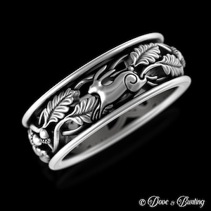 May include: A silver ring featuring an intricate design of a rabbit running through a leafy branch with acorns. The ring is a wide band with a raised design.