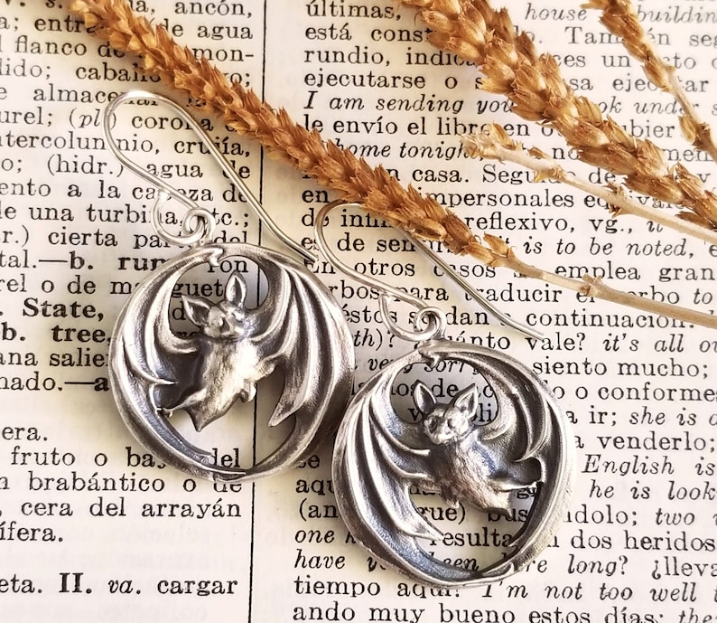 May include: A pair of silver earrings featuring a bat design. The bats are depicted in flight with their wings spread wide. The earrings are circular in shape and have a smooth, polished finish.