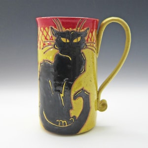 May include: A yellow ceramic mug with a black cat design. The cat is sitting with its tail curled around its body. The mug has a red rim and a handle.
