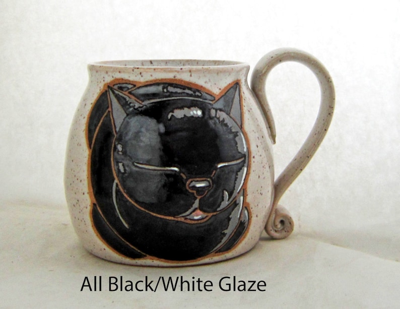 Pode incluir: Uma caneca de cer&acirc;mica com um desenho de gato preto. A caneca tem um exterior bege salpicado com uma ilustra&ccedil;&atilde;o de gato preto. A al&ccedil;a &eacute; bege e tem um design enrolado na base. O texto "All Black/White Glaze" est&aacute; na parte inferior.