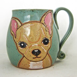 Puede incluir: Una taza de cer&aacute;mica con una imagen pintada a mano de un chihuahua marr&oacute;n y blanco con orejas rosas. La taza tiene un esmalte verde azulado y una asa.