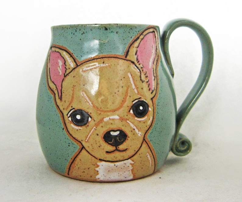 Puede incluir: Una taza de cer&aacute;mica con una imagen pintada a mano de un chihuahua marr&oacute;n y blanco con orejas rosas. La taza tiene un esmalte verde azulado y una asa.