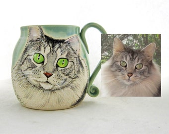 May include: A ceramic mug featuring a hand-painted portrait of a grey and white cat with green eyes. The cat is looking directly at the viewer. The mug has a light blue glaze and a handle shaped like a cat's tail.