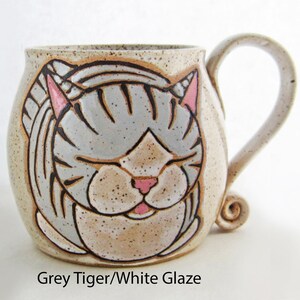 Pode incluir: Uma caneca de cer&acirc;mica com um desenho de rosto de gato. O gato tem listras cinzentas, orelhas e nariz rosa e olhos fechados. A caneca tem um acabamento bege salpicado e uma al&ccedil;a curva. O texto na parte inferior diz "Grey Tiger/White Glaze".
