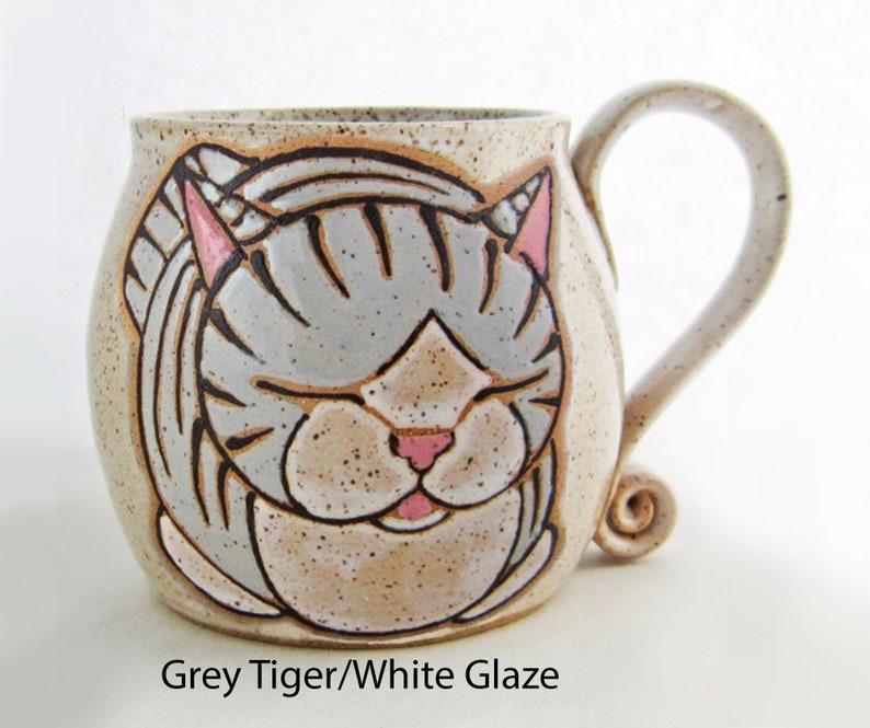 Pode incluir: Uma caneca de cer&acirc;mica com um desenho de rosto de gato. O gato tem listras cinzentas, orelhas e nariz rosa e olhos fechados. A caneca tem um acabamento bege salpicado e uma al&ccedil;a curva. O texto na parte inferior diz "Grey Tiger/White Glaze".