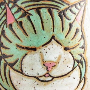 Pode incluir: Close-up de um rosto de gato de cer&acirc;mica com os olhos fechados. A pelagem do gato &eacute; verde claro com listras pretas, orelhas rosa e um nariz rosa. O fundo &eacute; branco salpicado.