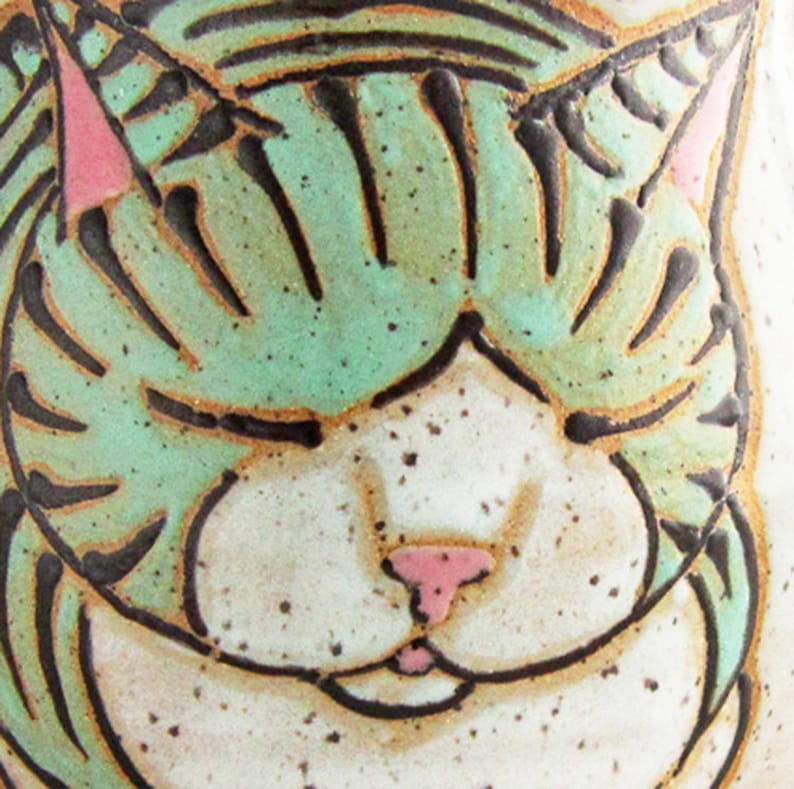 Pode incluir: Close-up de um rosto de gato de cer&acirc;mica com os olhos fechados. A pelagem do gato &eacute; verde claro com listras pretas, orelhas rosa e um nariz rosa. O fundo &eacute; branco salpicado.