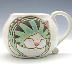 Könnte beinhalten: Ein weißer Keramikbecher mit bauchigem Körper und geschwungenem Henkel. Auf der Vorderseite befindet sich eine handgemalte Illustration einer schlafenden Katze mit mintgrünen und schwarzen Streifen, weißem Gesicht sowie kleinen rosa Ohren.