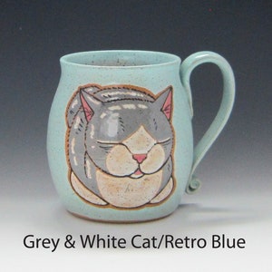 Pode incluir: Uma caneca de cer&acirc;mica em azul retr&ocirc; com uma ilustra&ccedil;&atilde;o pintada &agrave; m&atilde;o de um gato cinza e branco dormindo. A caneca tem formato arredondado, uma al&ccedil;a curva e uma textura salpicada. O texto "Grey & White Cat/Retro Blue" est&aacute; na parte inferior.
