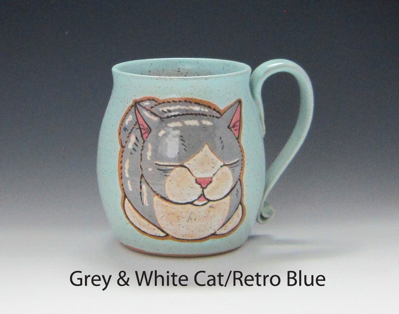 Pode incluir: Uma caneca de cer&acirc;mica em azul retr&ocirc; com uma ilustra&ccedil;&atilde;o pintada &agrave; m&atilde;o de um gato cinza e branco dormindo. A caneca tem formato arredondado, uma al&ccedil;a curva e uma textura salpicada. O texto "Grey & White Cat/Retro Blue" est&aacute; na parte inferior.