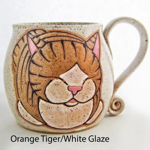 Pode incluir: Uma caneca de cer&acirc;mica com um desenho de rosto de tigre laranja pintado &agrave; m&atilde;o. A caneca tem um esmalte branco sujo salpicado e uma al&ccedil;a curva. O rosto do tigre apresenta listras marrons, orelhas e nariz rosa e um focinho branco. O texto "Orange Tiger/White Glaze" est&aacute; na parte inferior.