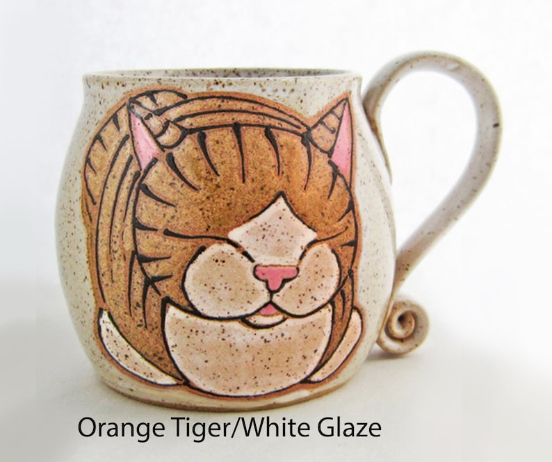 Pode incluir: Uma caneca de cer&acirc;mica com um desenho de rosto de tigre laranja pintado &agrave; m&atilde;o. A caneca tem um esmalte branco sujo salpicado e uma al&ccedil;a curva. O rosto do tigre apresenta listras marrons, orelhas e nariz rosa e um focinho branco. O texto "Orange Tiger/White Glaze" est&aacute; na parte inferior.