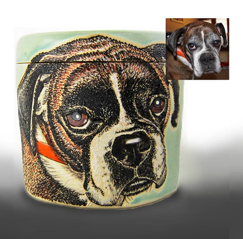 K&ouml;nnte beinhalten: Eine Keramikdose mit einer handgemalten Illustration eines Boxerhundes. Die Dose hat einen hellblauen Hintergrund und der Hund ist in Braun-, Schwarz- und Wei&szlig;t&ouml;nen dargestellt. Der Hund tr&auml;gt ein rotes Halsband.