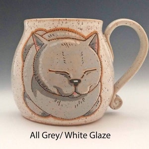 Pode incluir: Caneca de cer&acirc;mica com um desenho de rosto de gato cinzento sorridente. A caneca &eacute; branca suja com detalhes salpicados e uma al&ccedil;a curva. O texto "All Grey/ White Glaze" est&aacute; na parte inferior. Um item artesanal.