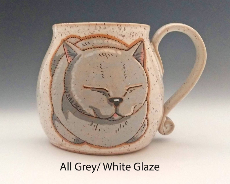 Pode incluir: Caneca de cer&acirc;mica com um desenho de rosto de gato cinzento sorridente. A caneca &eacute; branca suja com detalhes salpicados e uma al&ccedil;a curva. O texto "All Grey/ White Glaze" est&aacute; na parte inferior. Um item artesanal.