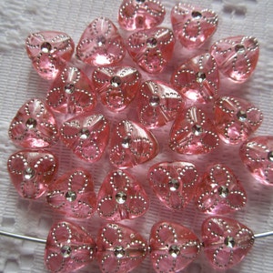Puede incluir: Una colección de cuentas de corazón rosa con detalles plateados y diamantes de imitación transparentes. Las cuentas están dispuestas en un montón suelto sobre una superficie blanca.