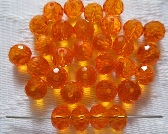 Puede incluir: Una colección de cuentas de cristal facetadas de color naranja. Las cuentas son redondas y tienen un agujero en el centro. Están dispuestas en una pila sobre una superficie blanca.