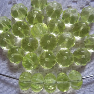 May include: A collection of 25 light green glass beads with a flower-like design. The beads are arranged in a loose pile on a white surface.