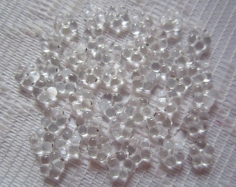 May include: A collection of clear, spherical water beads, clustered together on a white, textured surface. The beads have a translucent appearance, reflecting light and creating a sparkling effect. The image showcases the texture and form of the water beads.