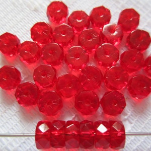 May include: A collection of faceted, transparent red beads. The beads are scattered across a white surface, with a strand of beads strung on a wire at the bottom. The beads have a geometric shape and are likely used for jewelry making.