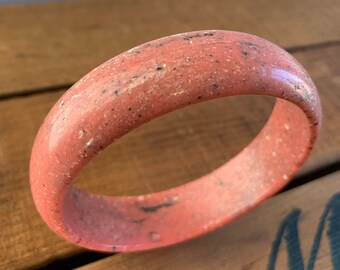 May include: A coral-colored bangle bracelet with a speckled texture. The smooth, rounded surface of the bracelet is a solid, warm hue with small, dark flecks. The bracelet is a simple, elegant accessory.