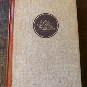 May include: A vintage book with a beige linen cover and a brown spine. A circular emblem featuring a house, trees, and a crescent moon is centered on the cover. The book's edges show signs of wear, suggesting its age.