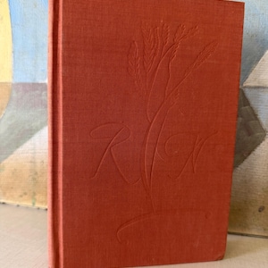 May include: A hardcover book with a solid burnt orange fabric cover. The front cover features an embossed design of wheat stalks and the stylized, intertwined capital letters R and W centered on the surface, creating a subtle textured appearance.