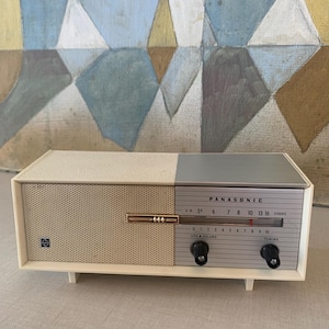 May include: Vintage Panasonic radio with a cream and gray color scheme. The radio features a speaker grille, tuning dial, and the brand name "PANASONIC". The radio has a retro design and is likely from the mid-20th century.