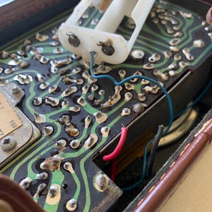 May include: Close-up of an open vintage electronic device. The interior reveals a circuit board with green pathways, metallic components, and wiring in blue, red, and black. A white plastic battery holder is visible.