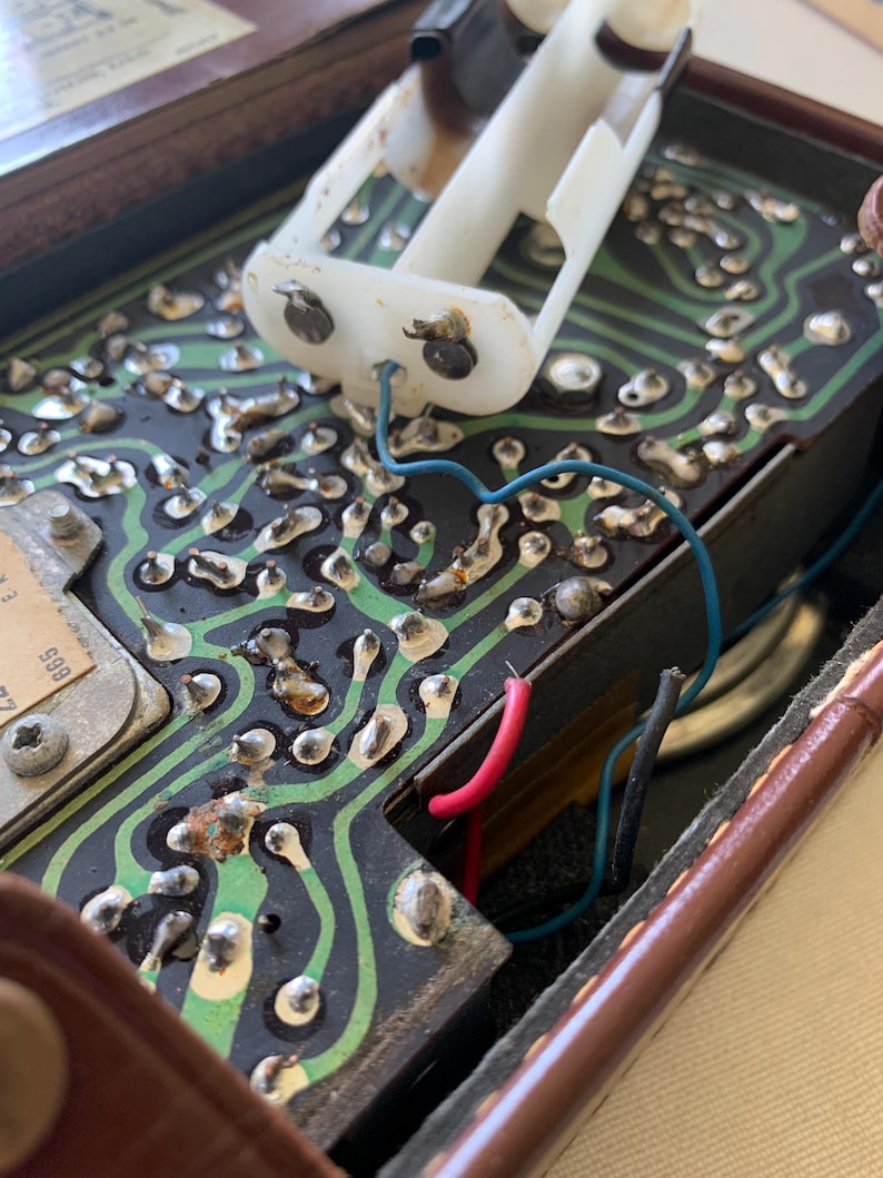 May include: Close-up of an open vintage electronic device. The interior reveals a circuit board with green pathways, metallic components, and wiring in blue, red, and black. A white plastic battery holder is visible.