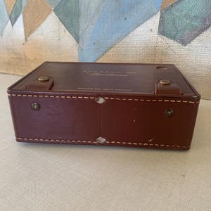 May include: A rectangular, brown leather case with the text "Emerson 888 Sportman" and "Genuine Top Grain Cowhide". The case has a stitched border and metal hardware. The case is closed.