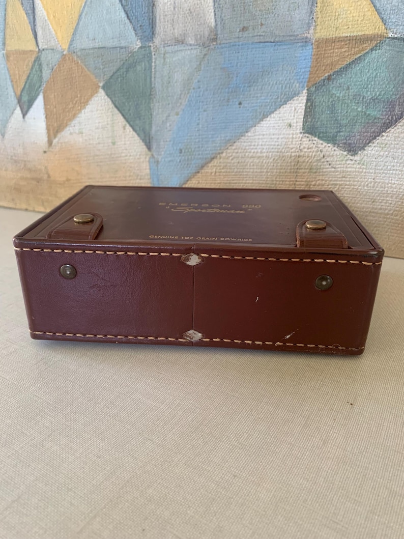 May include: A rectangular, brown leather case with the text "Emerson 888 Sportman" and "Genuine Top Grain Cowhide". The case has a stitched border and metal hardware. The case is closed.