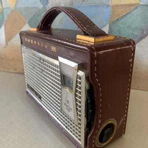 May include: Vintage Emerson 990 radio in a brown leather case with a handle. The radio has a silver metal speaker grill and a tuning dial. The text "Emerson 990" is printed on the front of the radio.