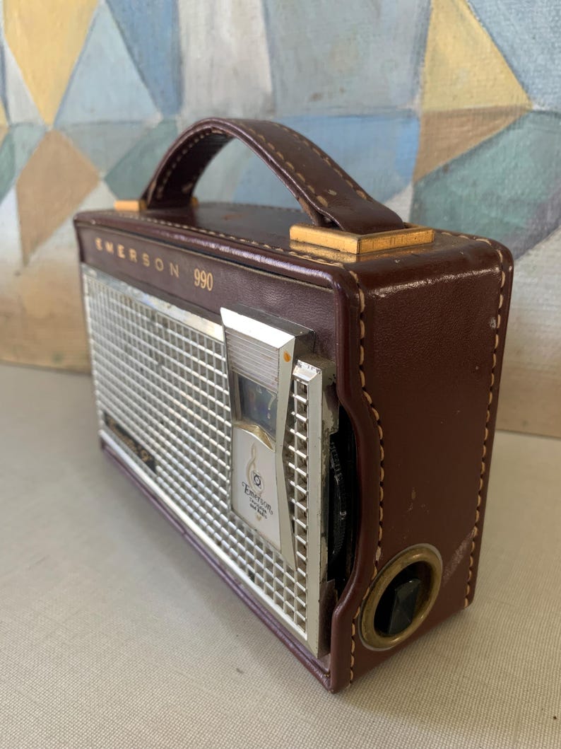 May include: Vintage Emerson 990 radio in a brown leather case with a handle. The radio has a silver metal speaker grill and a tuning dial. The text "Emerson 990" is printed on the front of the radio.