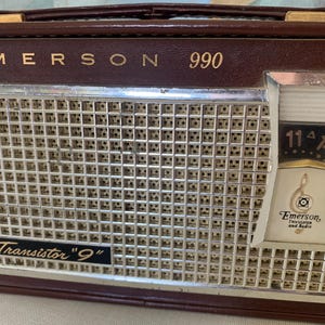 May include: Vintage Emerson 990 transistor radio in a brown leatherette case. The radio features a silver speaker grill, a tuning dial, and the text "Transistor '9'". The Emerson logo is visible on the dial.